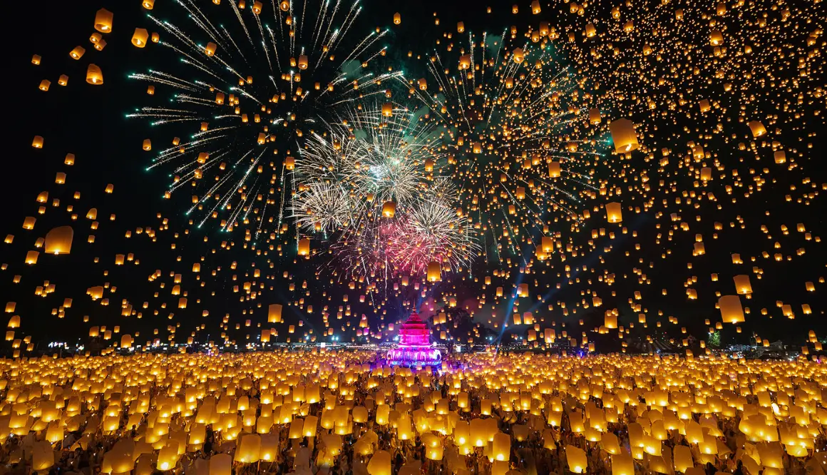 Milhares de lanternas douradas subindo ao céu noturno durante o Festival Yi Peng em Chiang Mai, refletidas na água
